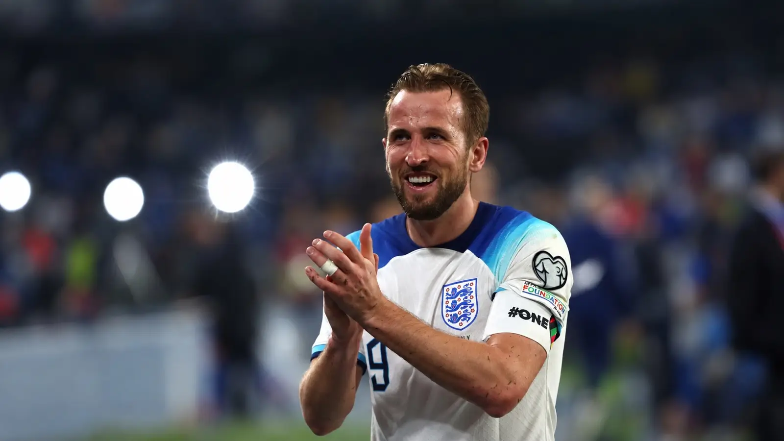 Harry Kane smiles and applauds supporters while wearing England’s white shirt and captain’s armband.