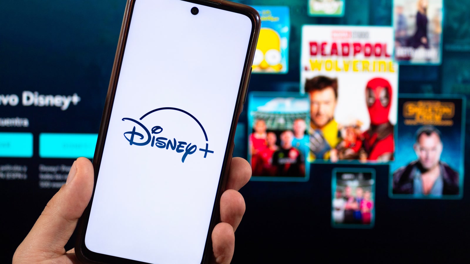 A hand holds a phone displaying the Disney+ logo in front of the streaming service interface.