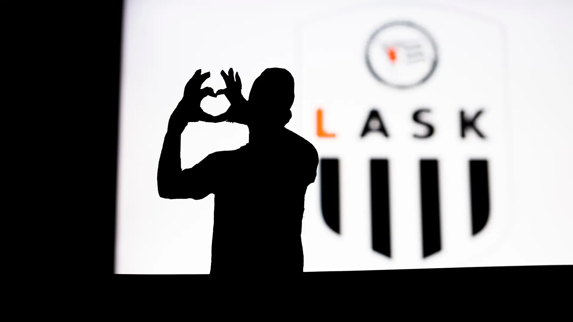 A silhouetted LASK supporter forms a heart with their hands in front of the club crest.