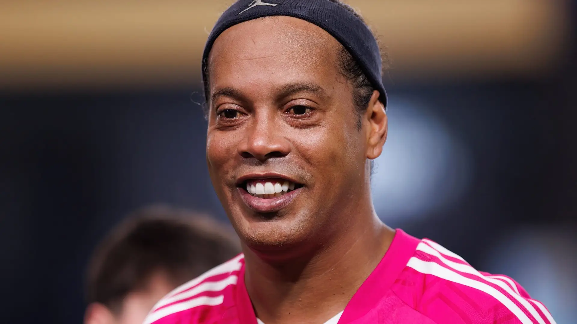 Ronaldinho smiling in a black cap and pink shirt.