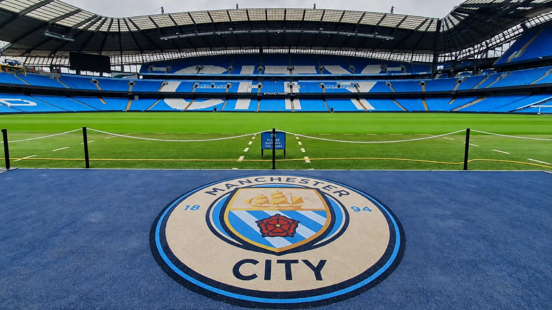 Manchester City crest beside the pitch at the Etihad Stadium, with blue seating in the background