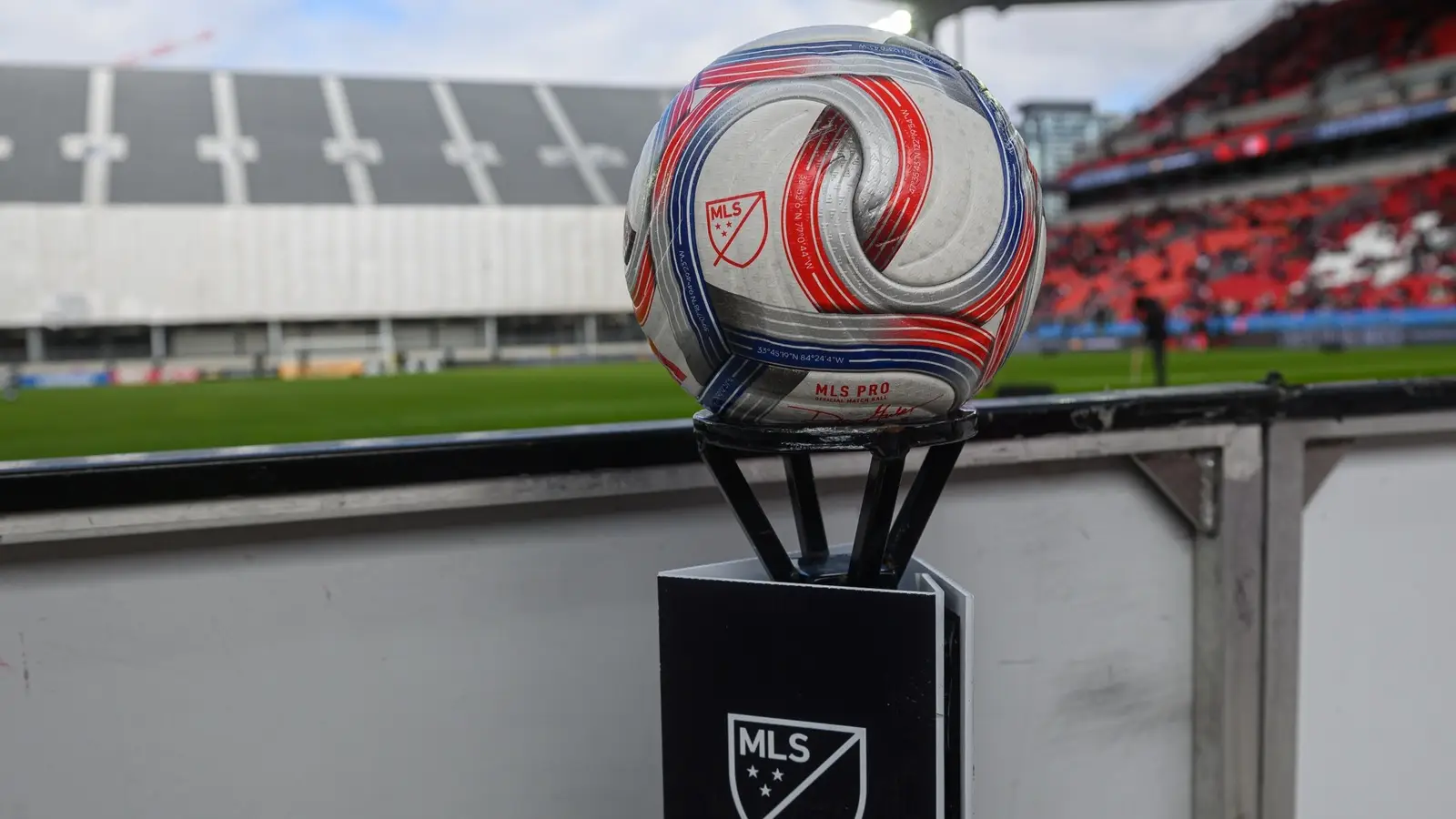 White, red and blue MLS match ball on a black MLS-branded pedestal beside a stadium pitch.