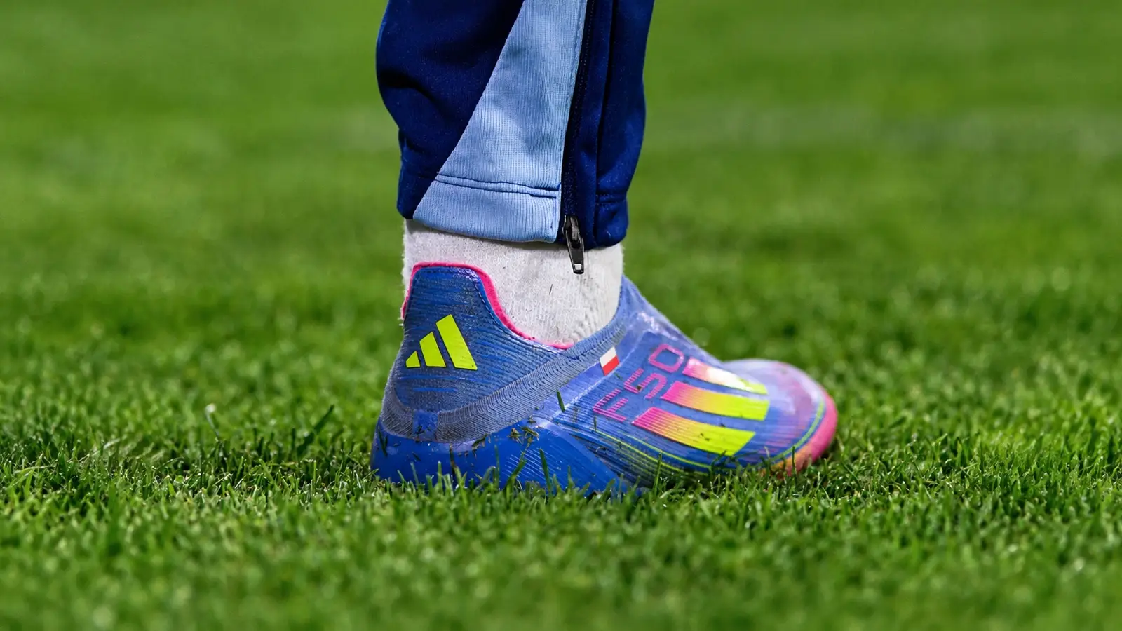 Blue Adidas F50 football boot with yellow and pink three stripes on a grass pitch