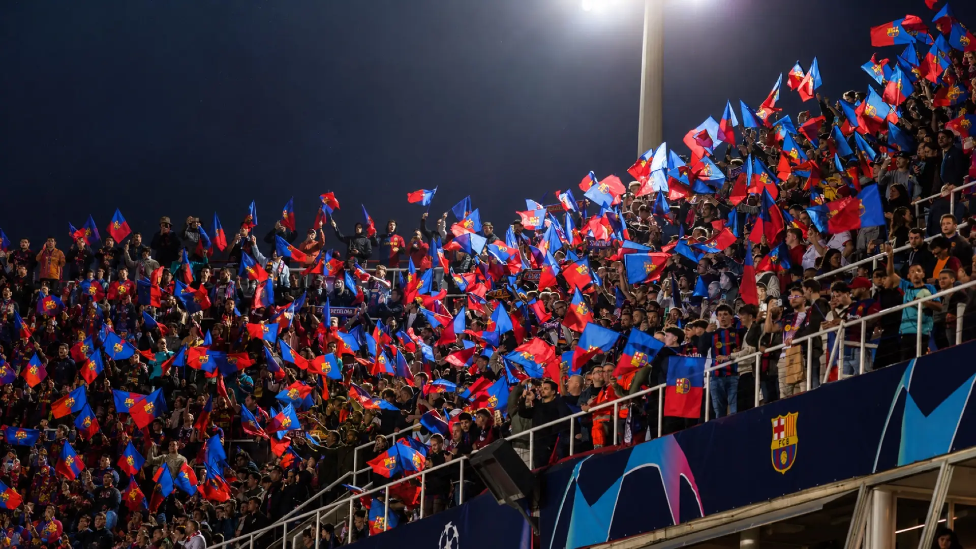 Barcelona supporters wave red and blue club flags in the stands under stadium floodlights.