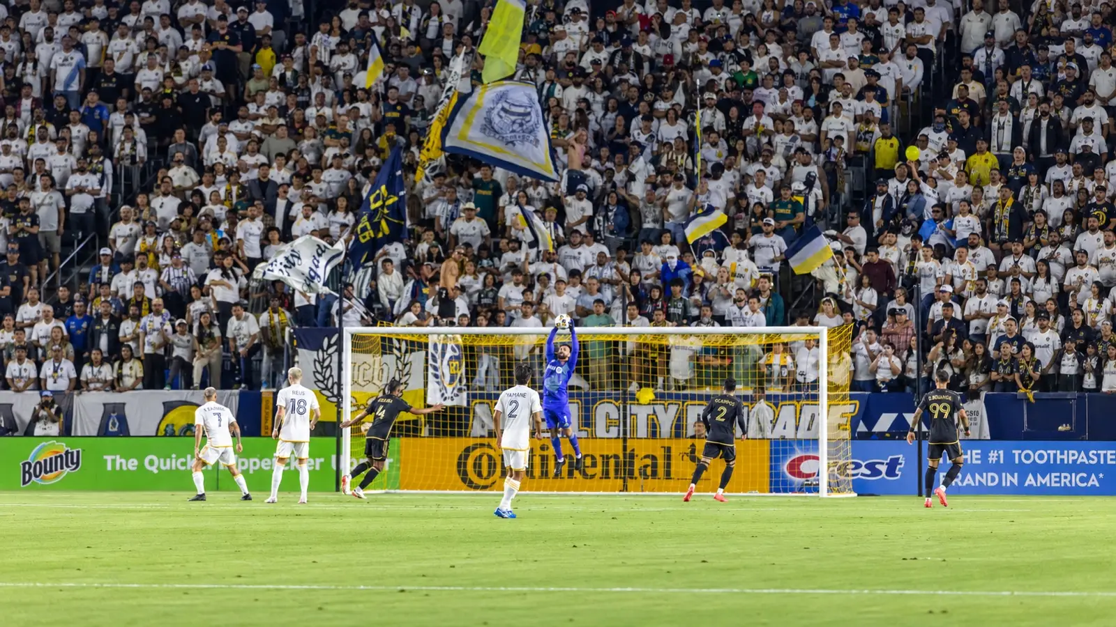 LA Galaxy goalkeeper catches the ball above LAFC and Galaxy players in front of flag-waving supporters during El Tráfico.