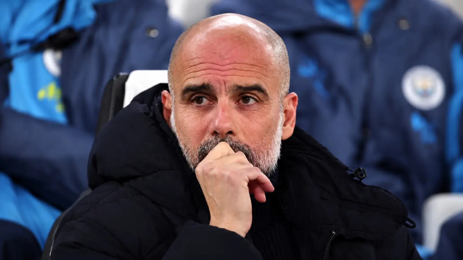Pep Guardiola seated in a stadium dugout with his hand near his mouth