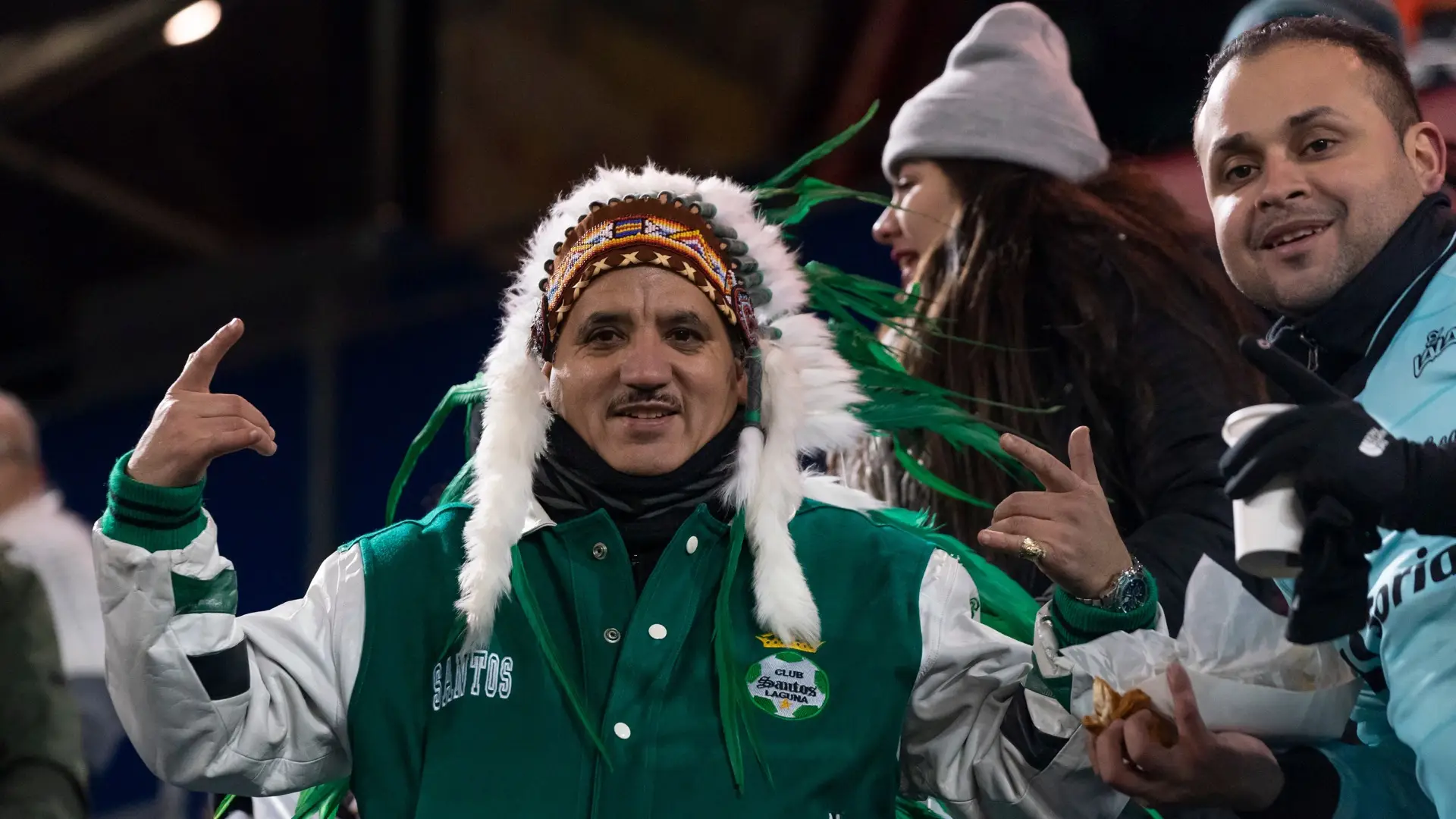 Santos Laguna supporter in a green club jacket and feathered headdress raises both hands in the stands.