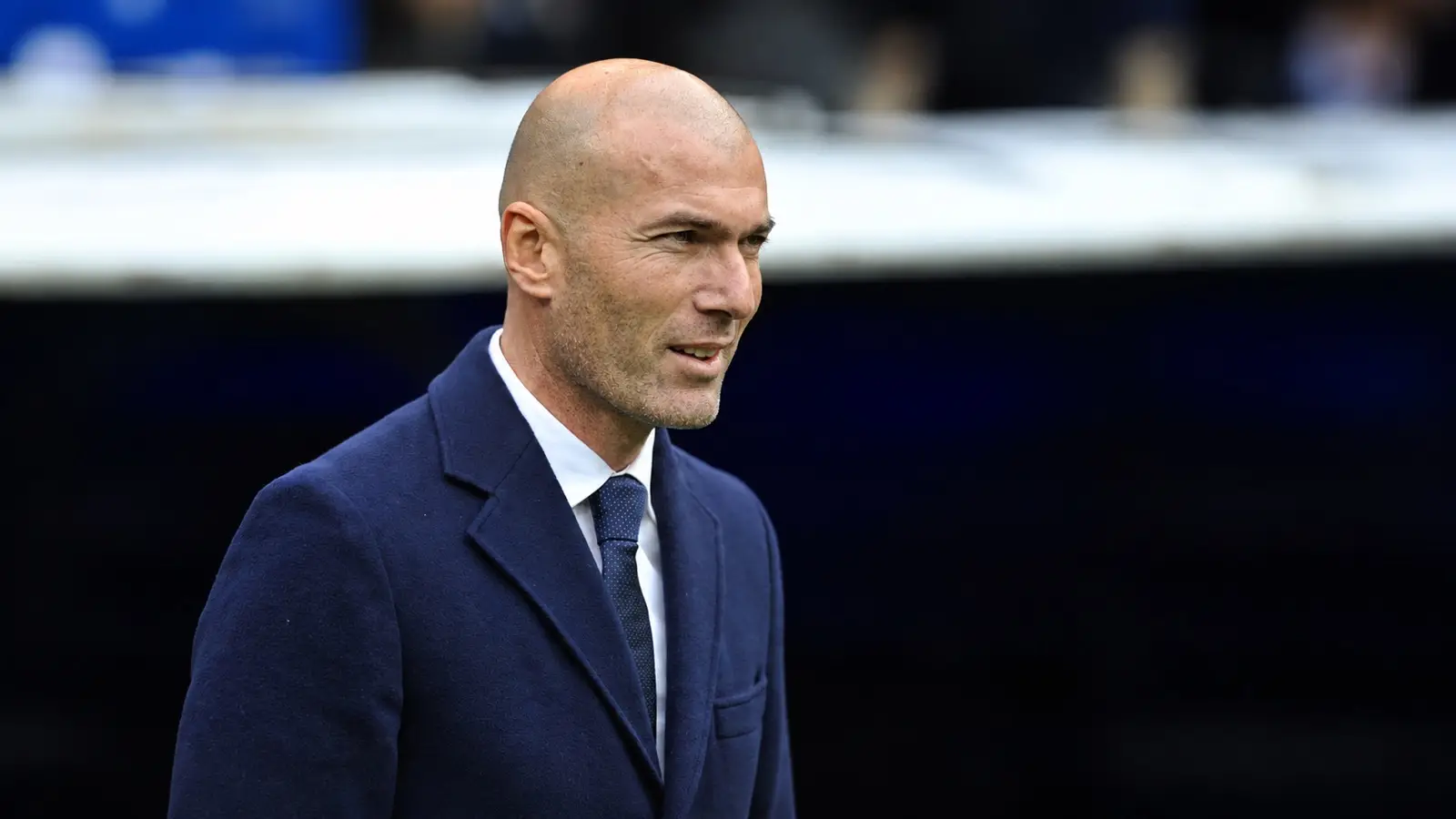 Zinédine Zidane in a navy coat and tie beside a football pitch